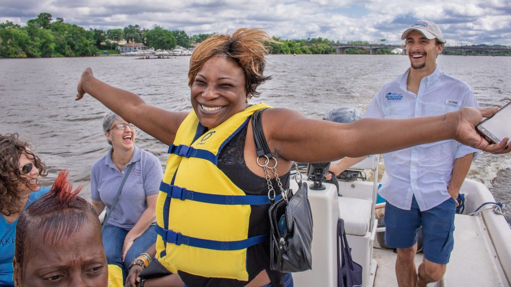 A woman in a lifejacket with outspread arms exuberantly enjoying the boat ride on the Anacostia.