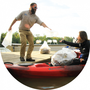Riverkeeper Trey Sherard reaching to give some more trash bags to a volunteer in a kayak with an already filled bag of trash.