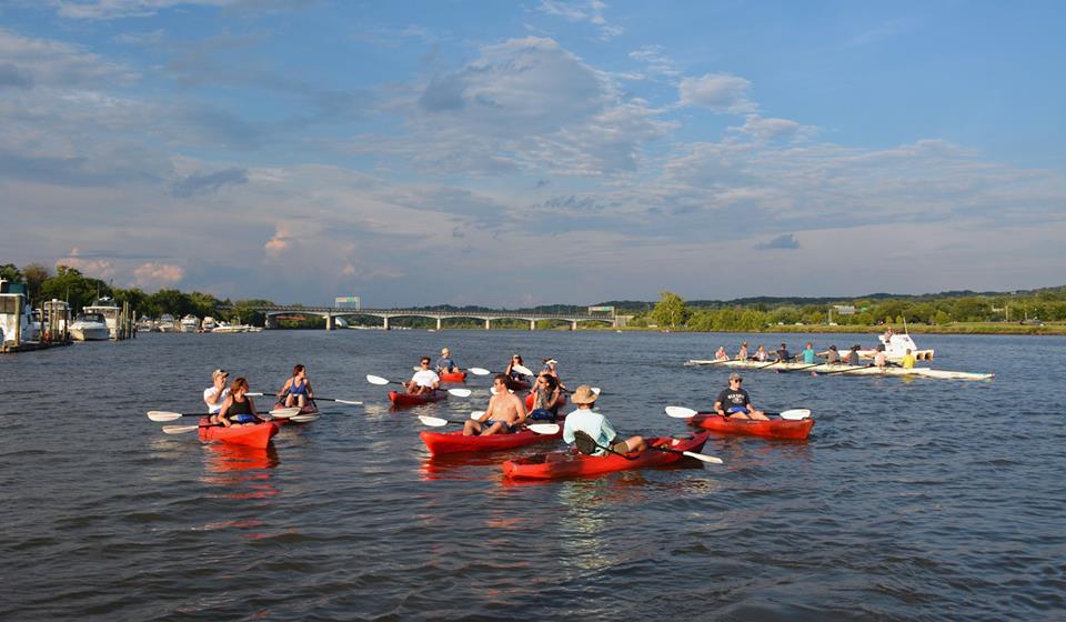 Recreation is growing on the Anacostia. A group of 9 kayaks in the water a the sun set, with an eight person racing scull in the background
